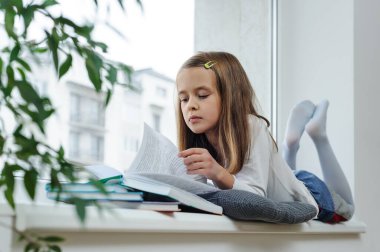 Kız pencere pervazında uzanıyor ve kitap okuyor..