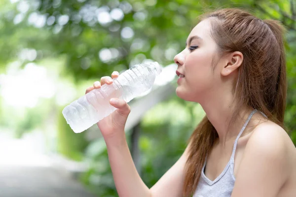 Portrait of young Asian woman drinking water bottle at summer green park.