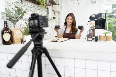 Çekici genç Asyalı güzel beyaz barista kahve dükkanı tezgahta video kamera çekimi kayıt ile gülümseyen önlük. Başlangıç küçük iş sahibi kavramı. Vlog kavram yapma.