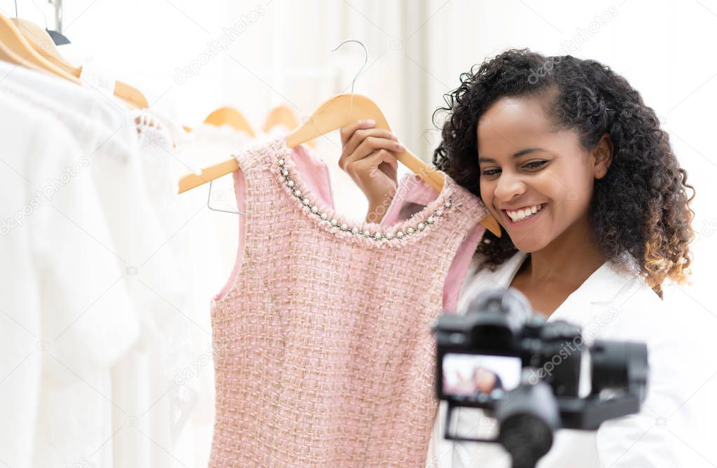 Young black smiling girl fashion stylish vlogger choosing apparel in clothing store holding with selfie video on the mirrorless Camera gimbal,recording video review on fashion trend, fashionista ,making vlog, vlog and social medial concept.