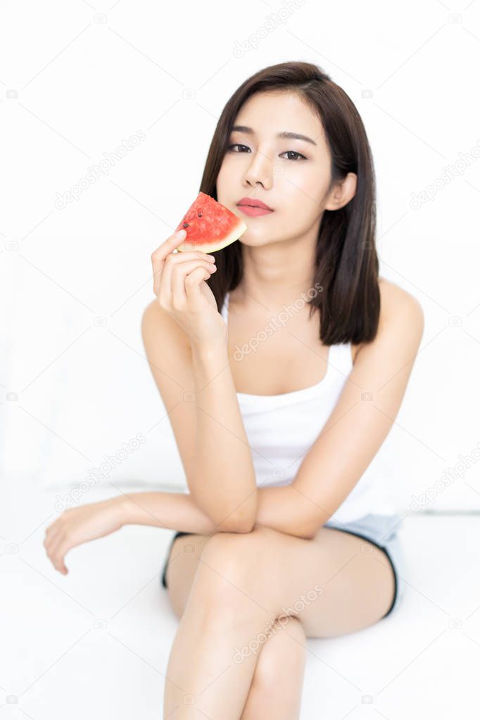 Portrait of a beautiful Asian young woman holding watermelon and looking at camera sitting on white sofa at her home (focus watermelon)
