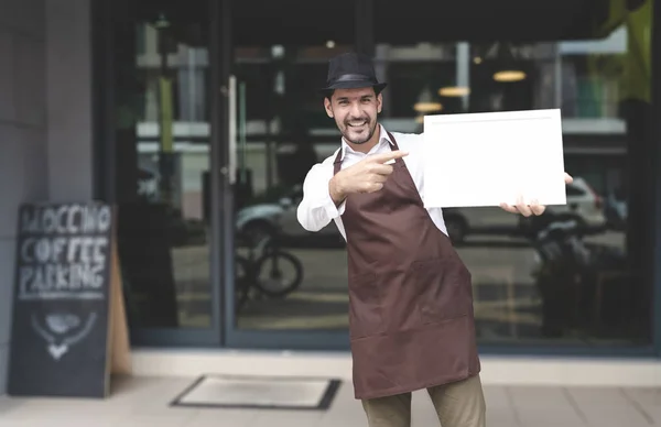 Portrait of smiling owner standing at front door of his coffee shop and pointing blank signboard. Young entrepreneur standing at the cafeteria door and looking at camera. Small Business owner concept.