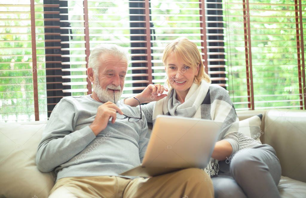 Happy senior couple looking at a laptop together on a sofa in their living room browsing the internet or social media with a smile