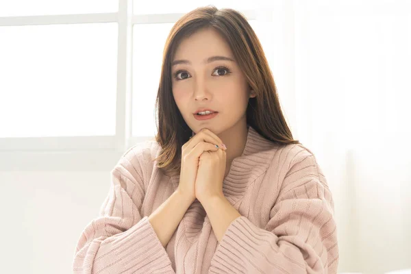 Portrait of inspired caucasian Asian lady wearing knitted sweater pink cold holding hands under chin and looking at camera in living room.Concept woman lifestyle and winter. Model fashion shooting. Autumn, winter season.