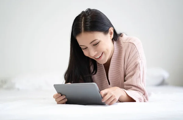 Happy young woman lying on the bed with tablet and surfing inter