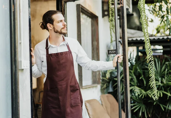 Красивый молодой человек в фартуке работает официанткой на открытии кафе — стоковое фото