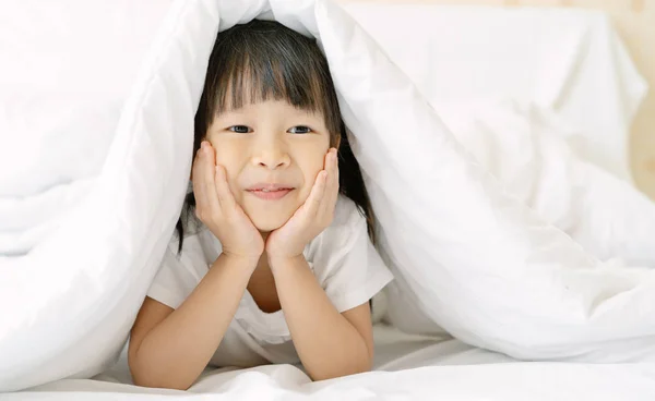 Nettes kleines Mädchen versteckt sich im Bett unter einer weißen Decke oder Decke — Stockfoto