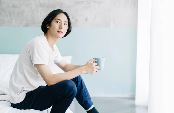 Porträtt av ung asiatisk man i pyjamas som håller kopp kaffe och — Stockfoto