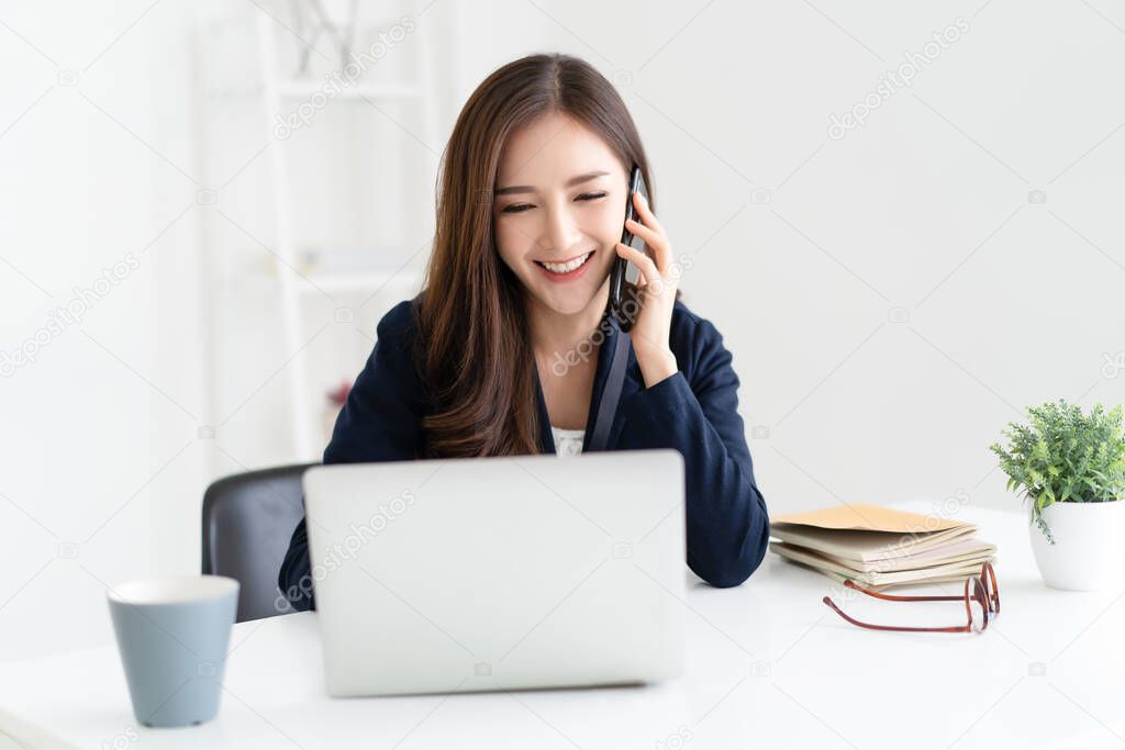 Smiling beautiful Asian woman freelancer using laptop working remotely from home. She is talking on the phone. Cozy office workplace, remote work, E-learning, New normal concept
