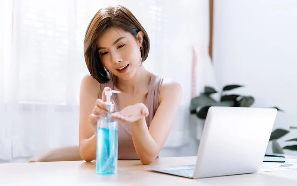 Portrait of beautiful Asian woman applying wash hand sanitizer gel bottle dispense for hands disinfection from bacteria while working from home, remote work. Hygiene and Covid-19 virus protection concept.