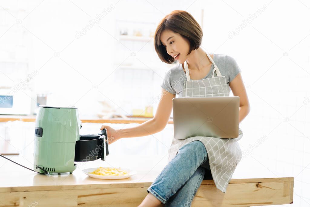 Portrait of beautiful Asian woman working on laptop while cooking with Air Fryers in kitchen during coronavirus pandemic.Remote work at home during self-isolation quarantine.New normal concept.