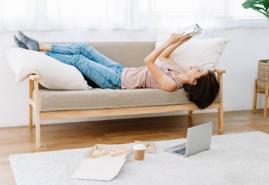 Güzel Asyalı kadın portresi kitap okuyor ve kanepeye uzanıyor, pizza yiyor ve Coronavirus salgınından kendini soyutlarken evde çalışıyor. Yeni normal yaşam tarzı konsepti..