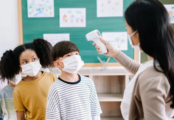 Multi-ethnic group of children with face mask stood in line let the teacher check, scan thermometer temperature for fever against the spread virus in classroom after covid-19 quarantine and lockdown.