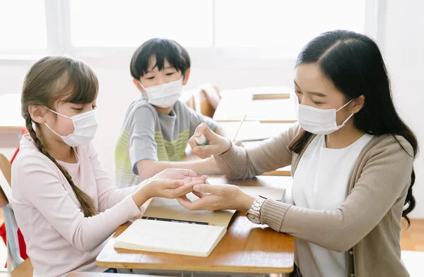 Female Asian teacher with protective face mask and sanitizing school children\'s hands with disinfectant spray in classroom after reopening school,covid-19 quarantine and lockdown.Healthcare concept.