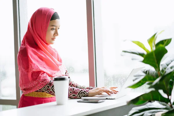 Portrait of young Muslim woman in hijab pink dress using laptop computer sitting at cafe table. Muslim business professional concept