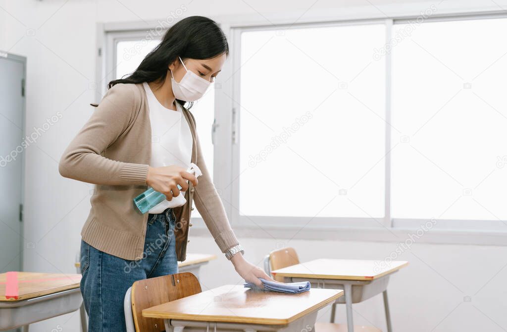 Young Asian female Teacher in a medical mask cleans up the desk in the classroom before students return to school after covid-19 quarantine and lockdown. Healthcare of pupils and students concept.