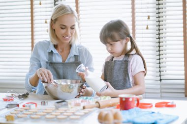 Anne ve kızı mutfakta birlikte yemek pişiriyorlar. Anne büyük bir karıştırma kabında hamuru karıştırırken kız bir şişe süt tutuyor, takım çalışması, öğrenme ve aile bağlarını gösteriyor..
