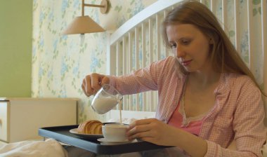 Genç bir kadın yatakta kahvaltı yapıyor. Bir fincan kahveye süt döker.