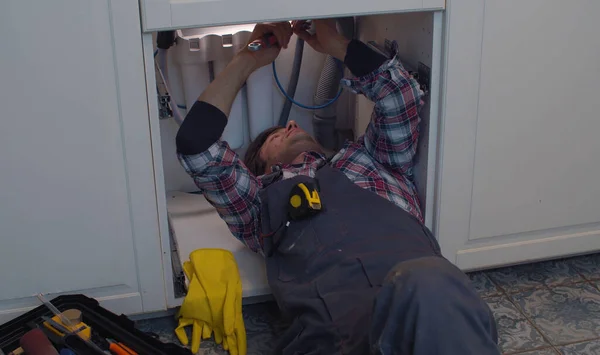 Plumber repairs a siphon leak in a kitchen sink. Young handsome worker lying on the floor and unscrewing the siphon detail in the kitchen cabinet. Minor repairs and maintenance of the home.