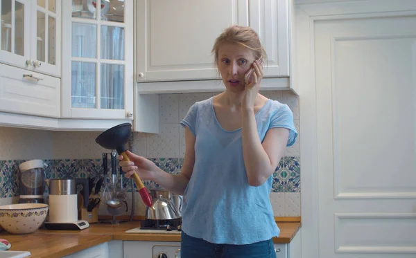 在厨房里困惑的女人手里拿着一个柱塞她正在用智能手机打电话寻求建议 厨房的水槽堵塞了 水管道需要修理 家政服务 免版税图库图片