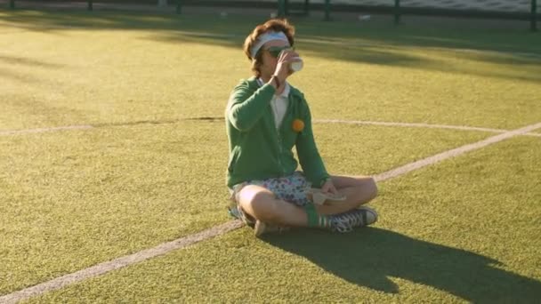 Funny freak sitting on the tennis court and drinking tea — Stock Video