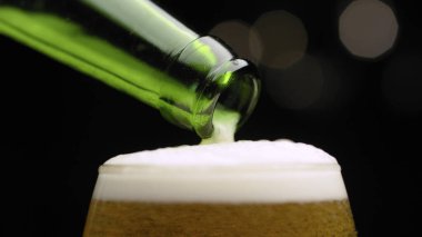 Koyu arkaplanda bir bardağa dökülen altın birayı kapat. Köpüklü su yeşil bir şişeden akıyor.