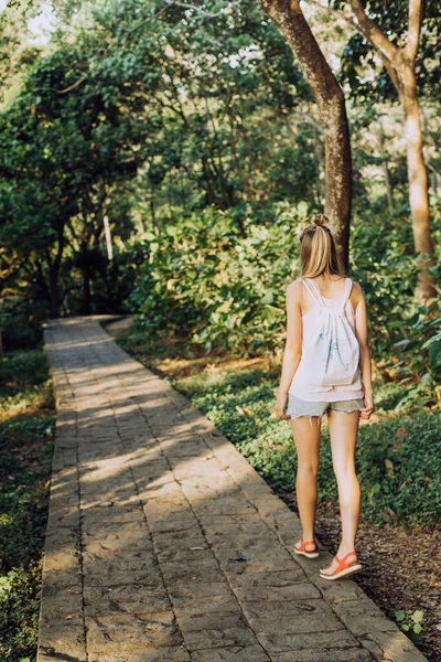 身着夏装背着背包的女孩走在丛林和茂密的绿树之间的石路边 探险及旅游 — 图库照片
