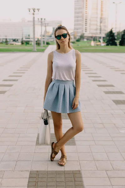 Fashionable girl in summer light clothes and glasses stands against the backdrop of the city. She is serious and looking at the camera.