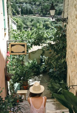 Sırtı açık şapkalı kız merdivenlerde oturuyor eski evlerle saksıdaki yeşil bitkiler arasında..