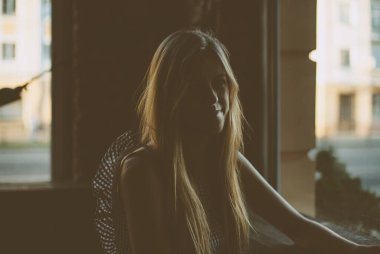 Uzun saçlı ve olağandışı elbiseli bir kız silueti. Karanlık bir odada oturuyor, utanarak gülümsüyor ve kameraya bakıyor..