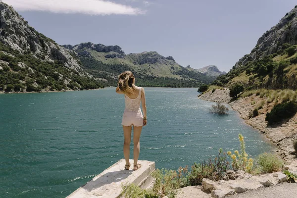 Traveling girl in summer clothes stands with her back to the camera, against the background of an azure mountain lake and green mountains.