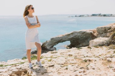 Açık renkli klasik elbiseli ve spor ayakkabılı Avrupalı bir kadın deniz kenarındaki kayanın üzerinde duruyor. Güneş gözlüğü takmış, başka tarafa bakıyor. Sağdaki imza için bir yer..