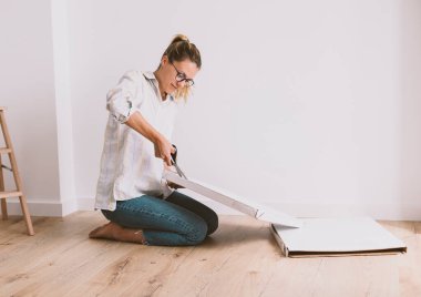 Gözlüklü genç bir kadın ahşap zeminde oturur ve beyaz kutuyu keser. Parlak bir oda, güzel bir ev..