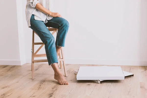 Woman in jeans and shirt sits on  wooden stepladder near white wall. Cardboard boxes are on the floor