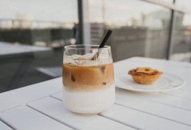 Beyaz masada buzlu latte ve siyah pipet. Yanında da börek tabağı var..