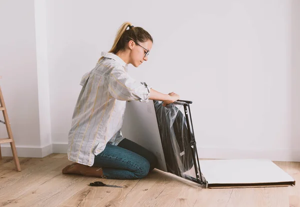 Pretty woman in work clothes sits on the floor in bright room and unpacks furniture.