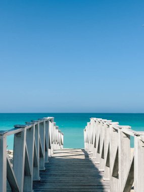 Okyanus ufkuna doğru beyaz tahta bir tahta kaldırımdan aşağı doğru simetrik bir manzara. Sakin, turkuaz su açık mavi gökyüzü ile buluşur, sakin ve davetkar bir tropik cennete giden yolu yaratır..