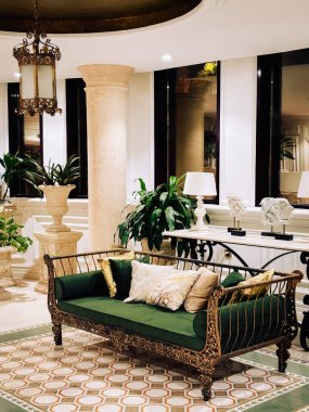 Lüks bir otel lobisinin ön planda büyük yeşil ve altın kadife bir kanepesi olan dekoratif bir iç çekimi. Dekoratif döşeme döşemesi, geniş bir sütun ve dekoratif ekicilerde yemyeşil bitkiler yer alıyor..