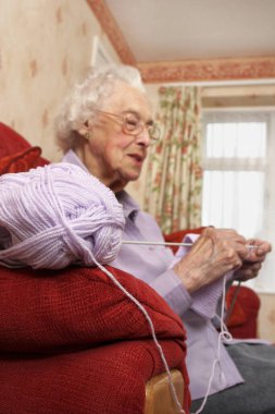 Kanepede oturmuş örgü ören gözlüklü yaşlı bir kadının yan görüntüsü.