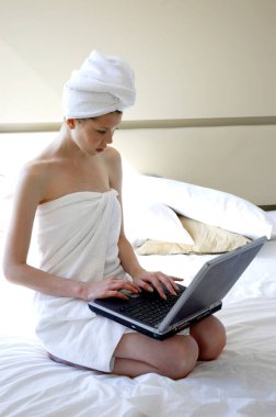 Bedeni ve saçı havluya sarılı bir kadın dizüstü bilgisayar kullanarak yatakta oturuyor.