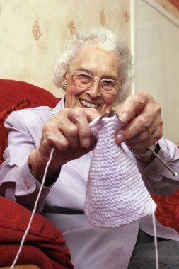 Kanepede oturan gözlüklü yaşlı bir kadın kameraya nasıl örgü ördüğünü gösteriyor.