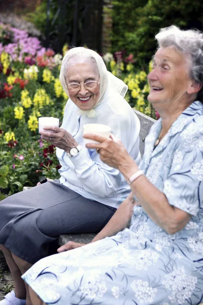 Two old women sitting on a bench in the park drinking coffee