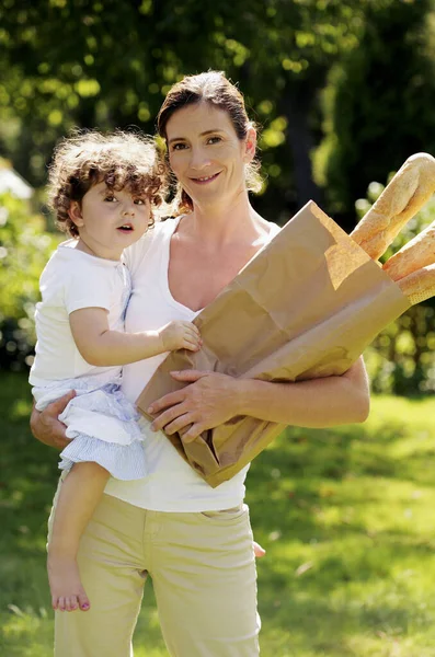 Woman holding a paper bag of french breads while carrying her daughter