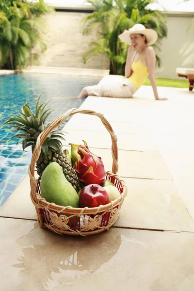 A basket of fruits with woman sitting by the poolside in the background