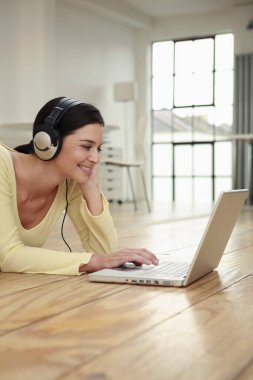 Kulaklıklı kadın dizüstü bilgisayar kullanarak yerde yatıyor.