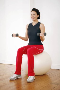 Bir kadın spor topunun üzerinde otururken halterleri kaldırıyor.