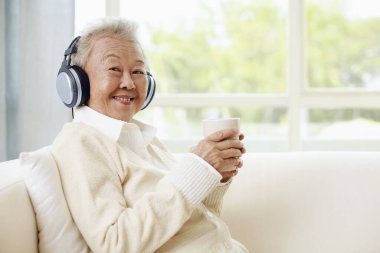 Kulaklıklı yaşlı kadın elinde bir fincan kahve tutuyor.