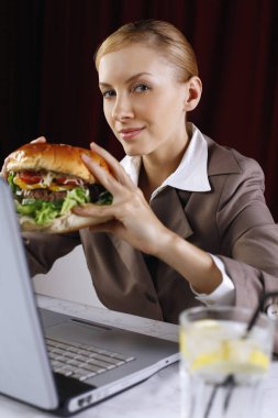 Masasında dizüstü bilgisayar kullanırken elinde hamburger tutan iş kadını.