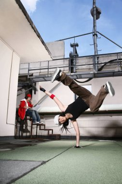 Breakdance hareketi yapan genç bir kadın, kulaklıklı genç bir adam arka planda dizüstü bilgisayar kullanıyor.