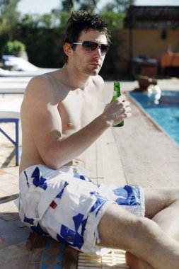Havuz kenarında oturan adam elinde bir şişe bira tutuyor.